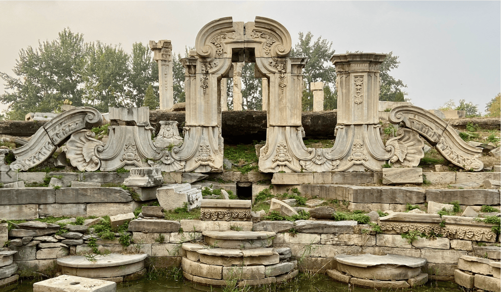Ruins of ornate stone structures with intricate carvings, surrounded by greenery and remnants of columns.