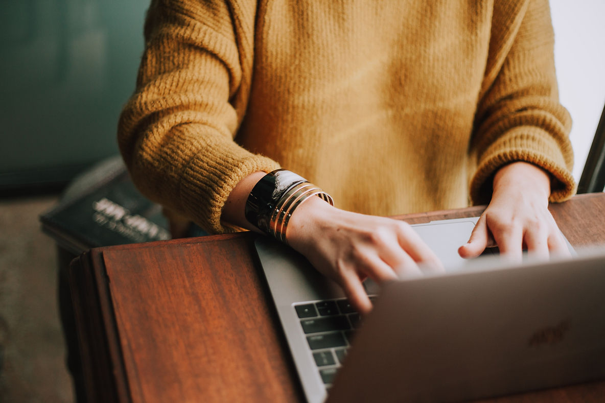 A person wearing a yellow sweater is typing on a laptop, showcasing their hand adorned with several bracelets. A stack of books is visible in the background.