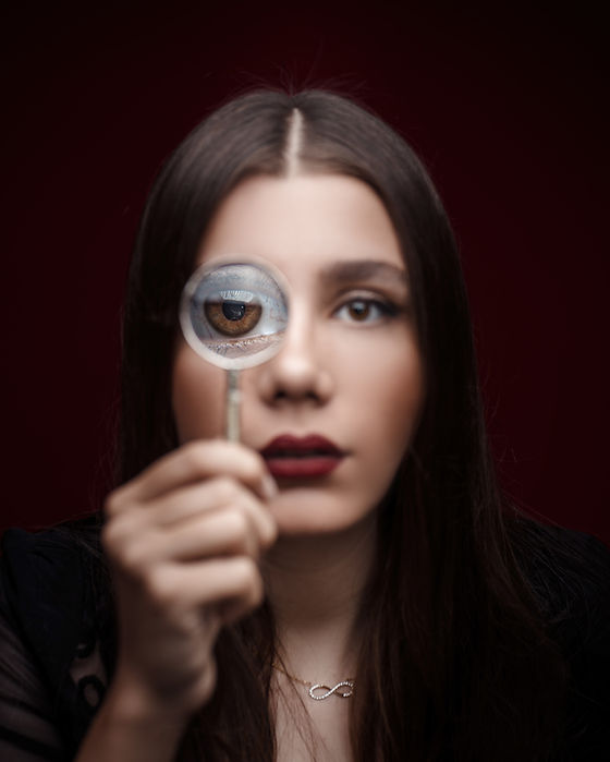 A young woman holding a magnifying glass to her eye, with a creative focus on her eye reflected in the glass. The background is a deep red, enhancing the dramatic effect.