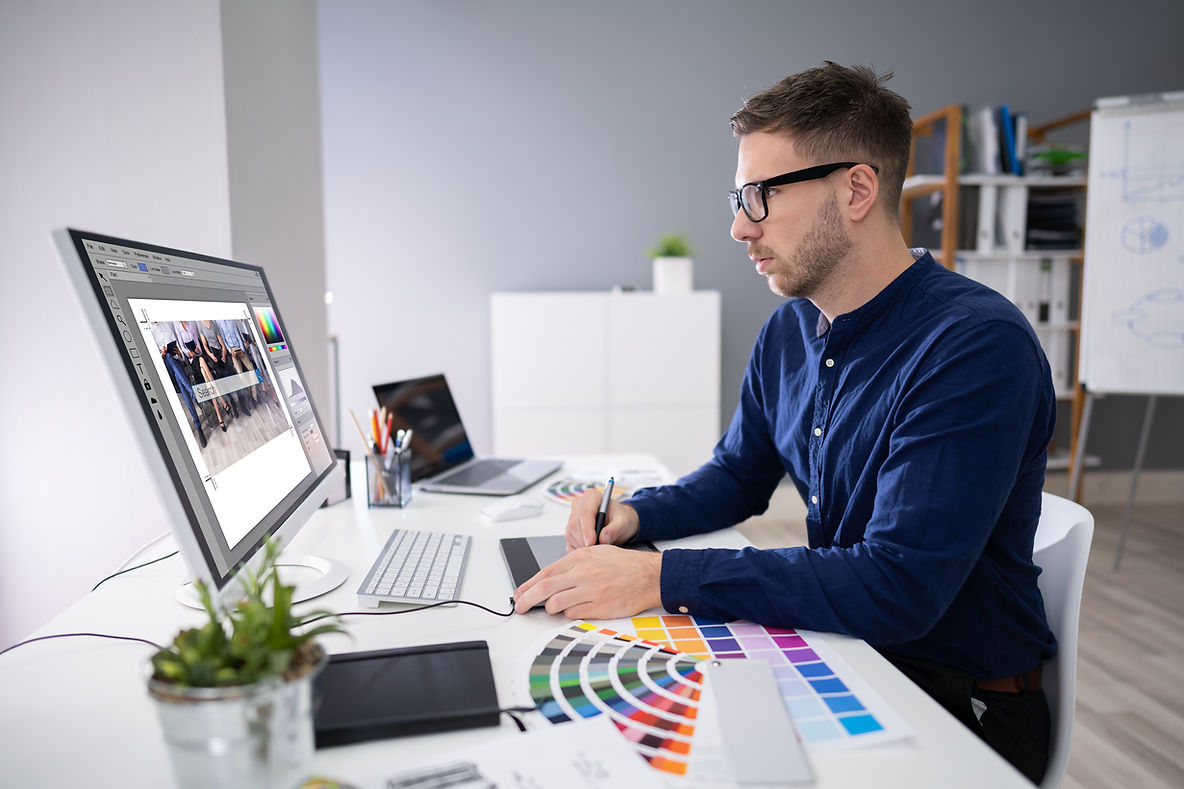 A man working at a desk with a computer, using a drawing tablet and surrounded by color swatches and design tools in a modern office setting.