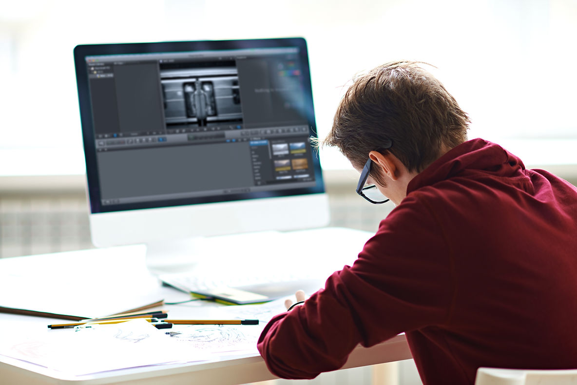 A person wearing glasses sits at a desk, focused on drawing on paper while a computer screen displays video editing software.