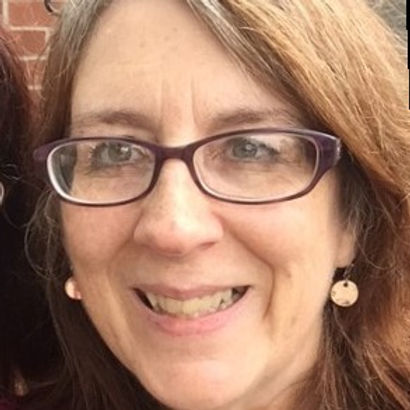 Close-up portrait of a woman with long, wavy brown hair, wearing purple glasses and dangling earrings, smiling.