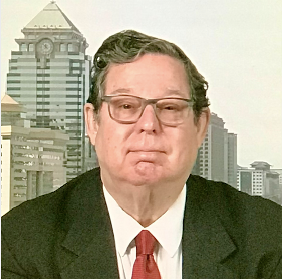 A man with glasses and a suit, sitting in front of a city skyline background.